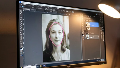 Close-up of a desktop monitor displaying the GIMP photo editor on Windows, with a portrait photograph open in the canvas and a layers panel on the right showing two stacked layers labeled "face_source" and "background". A free-select lasso outline traces a face on the upper layer, with a soft mask thumbnail visible. Cool desk lamp light from the upper right hits the matte screen, no glare, slight monitor reflection of warm room ambience. Style: technical screenshot photography. Mood: focused, hands-on.