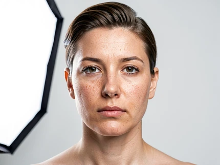 A close-up portrait of a person in a bright studio setting, face perfectly lit by a large softbox from the left, skin tones rendered with fine pore detail and natural subsurface scattering. The expression is neutral and composed, hair neatly styled. Background is a seamless light grey. Style: high-resolution commercial portrait photography. Mood: clean and lifelike.