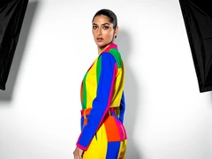 A person in a bold primary-color outfit stands against a clean white studio backdrop, posed with one shoulder forward and a direct gaze into the lens. Studio softbox lighting from both sides eliminates harsh shadows and renders skin tones evenly. Subtle gradient shadow falls behind the figure. Style: high-fashion editorial photography. Mood: confident and graphic.