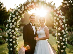 A couple in formal wedding attire stands beneath a floral arch of white roses and greenery in a sunlit garden, both smiling warmly at the camera. Golden-hour sunlight streams from behind, creating a soft rim light around their silhouettes and warm lens flare. Shallow depth of field blurs the garden path behind them. Style: romantic wedding editorial photography. Mood: joyful and intimate.